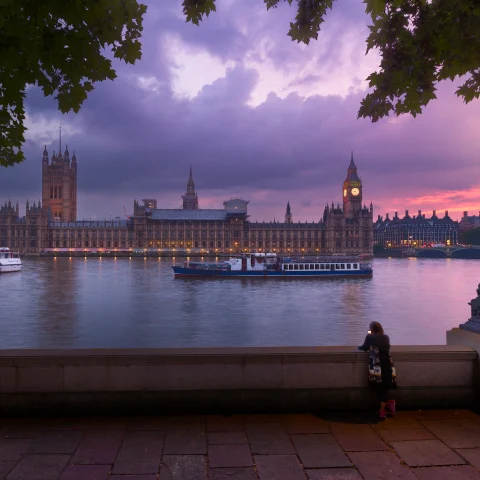 Parliament in London, England at sunset with pink and purple dark clouds and a woman standing across the river next to a lamp post.