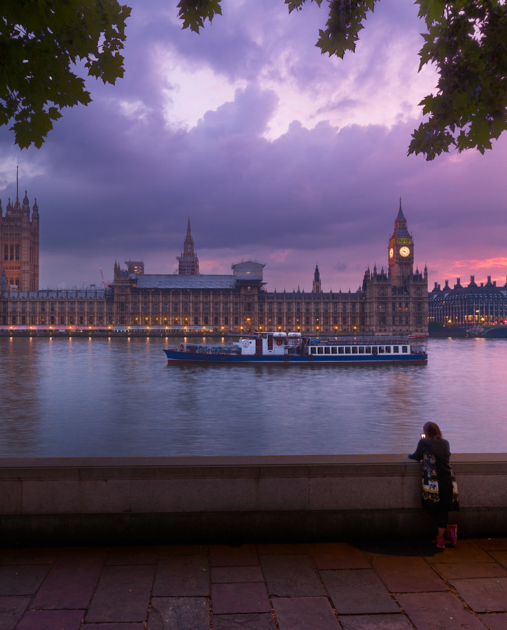 Parliament in London, England at sunset with pink and purple dark clouds and a woman standing across the river next to a lamp post.