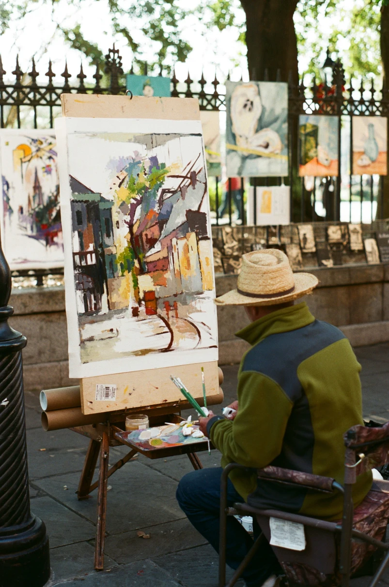 A man wearing a straw hat and green jacket, sitting on a stool and painting a picture outside. 