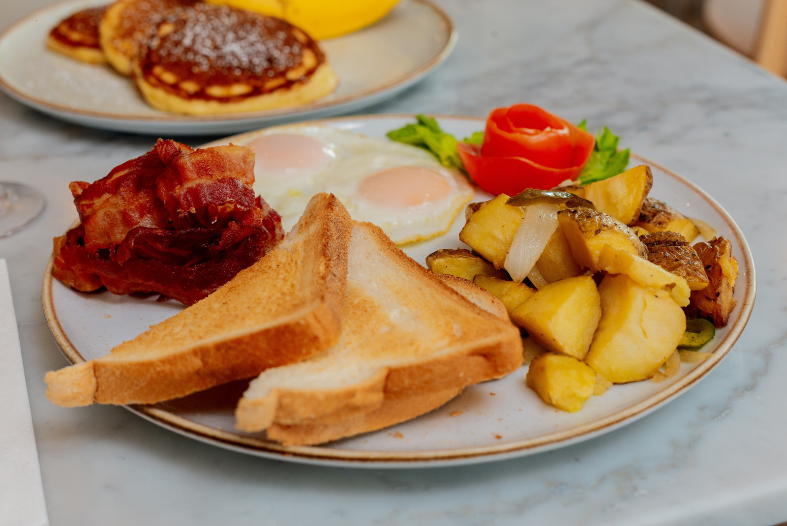 Toast, bacon, eggs and potatoes on a white plate and plate of pancakes in the background