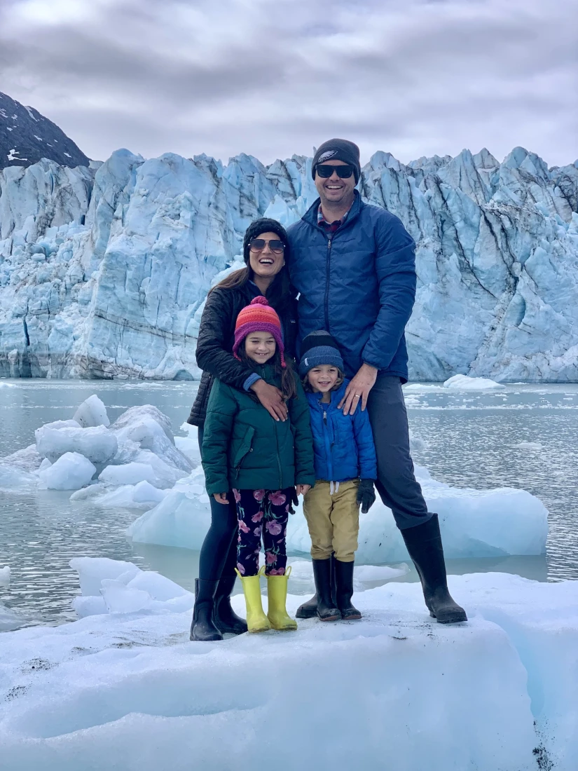 a man, a woman, and two small children in winter coats stand in front of a towering glacier