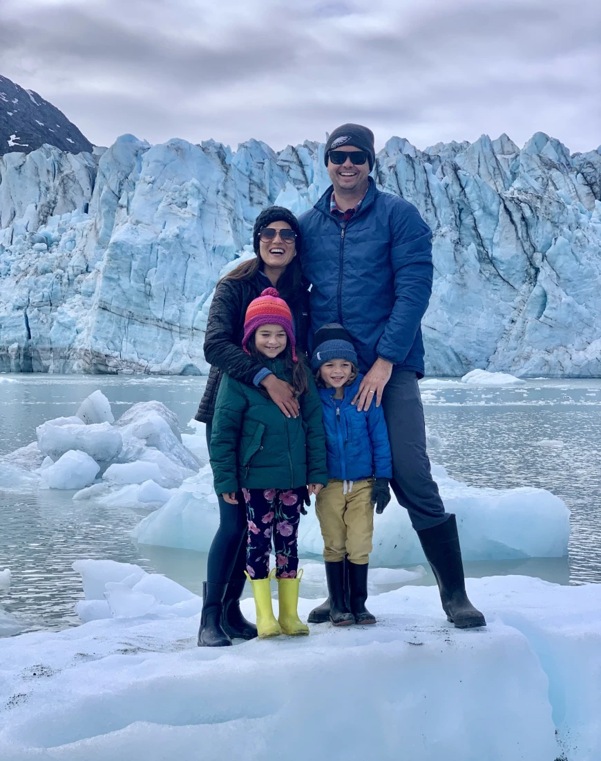 a man, a woman, and two small children in winter coats stand in front of a towering glacier