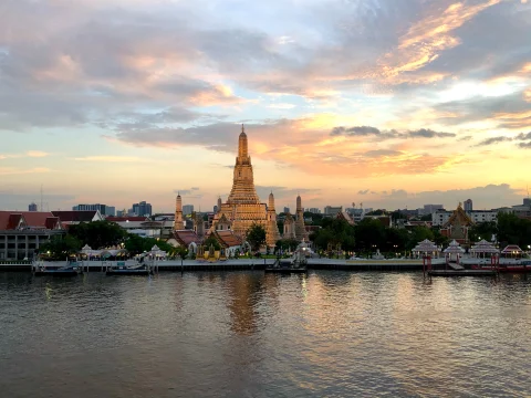A yellow and pink sunset over a golden temple in front of a still, dark blue river in Bangkok, Thailand.