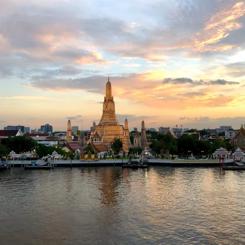 A yellow and pink sunset over a golden temple in front of a still, dark blue river in Bangkok, Thailand.