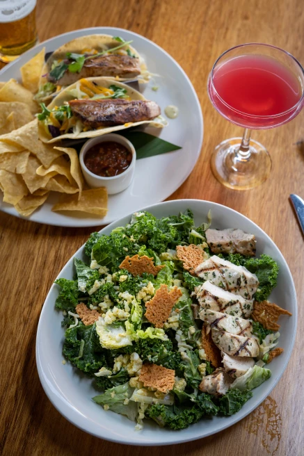 Fish tacos and a green kale salad on white plates with a pink cocktail at Leilani's On The Beach in Maui, Hawaii.