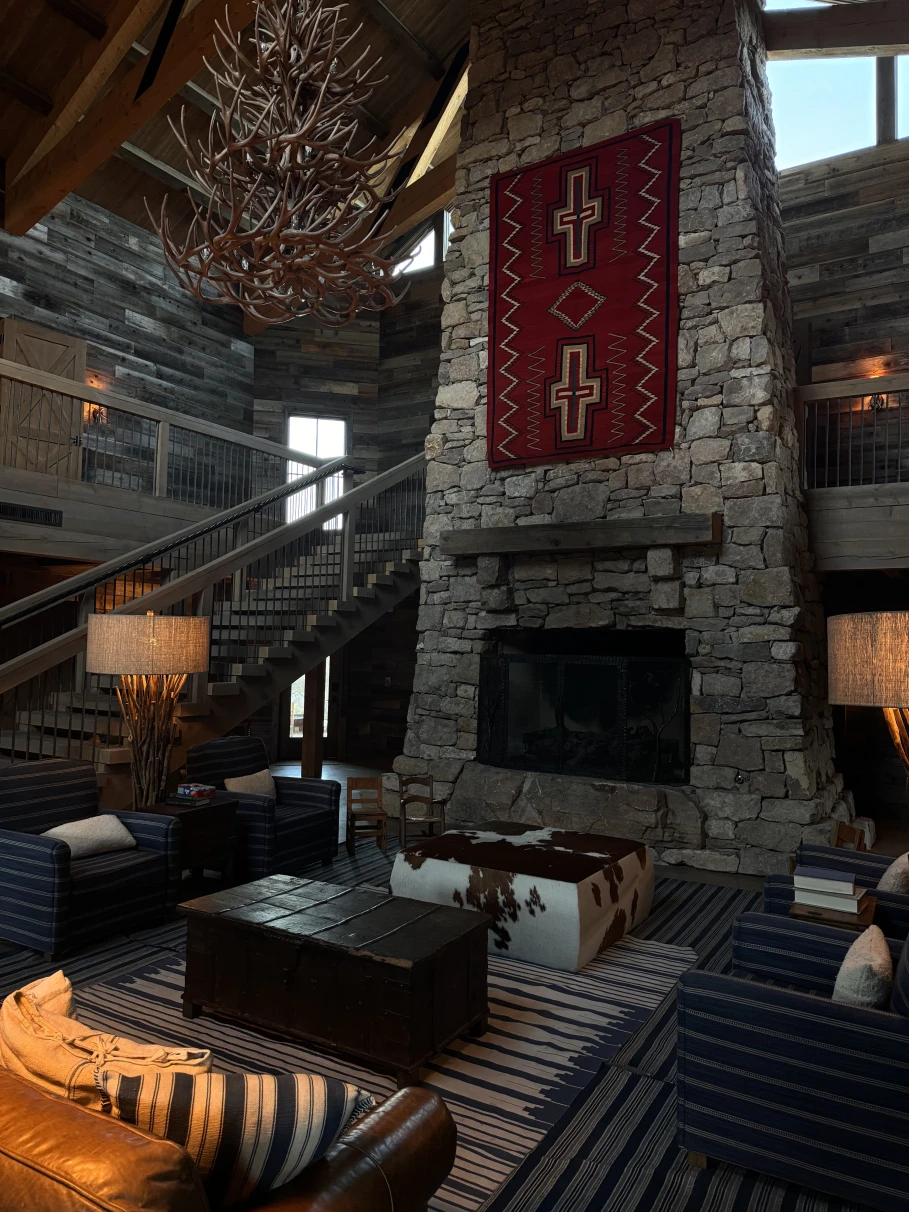 An image of a stone fireplace with a red carpet, wooden chandelier, seating area, leather couch, stairwell and carpet. 