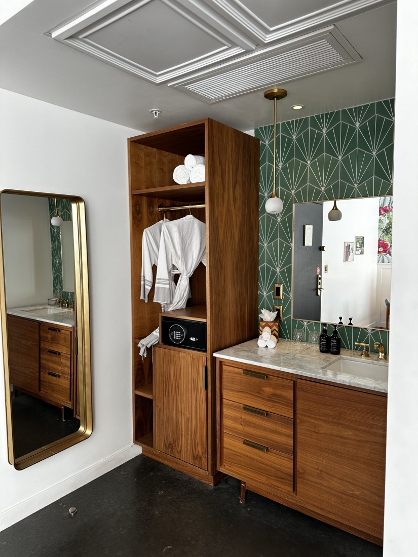 A hotel bathroom complete with two mirrors, patterned wall paper, hanging pendants, robes hanging inside of a closet, and cabinetry. 