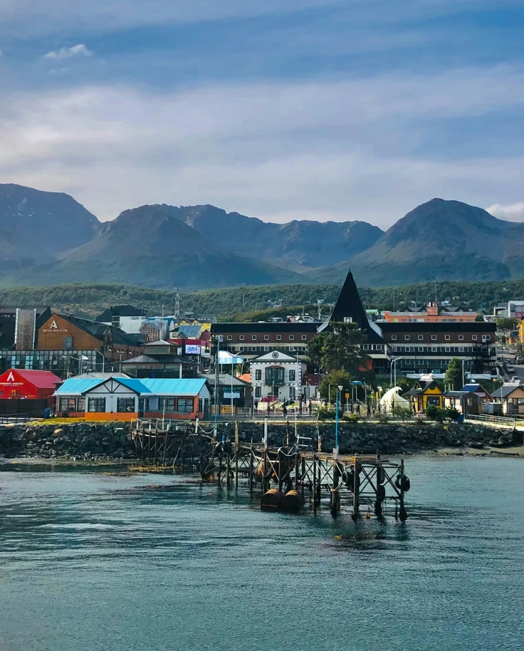 A blue lake with pink, blue and yellow houses and boats with green and brown mountains in the distance in Ushuaia on a Argentina vacation.