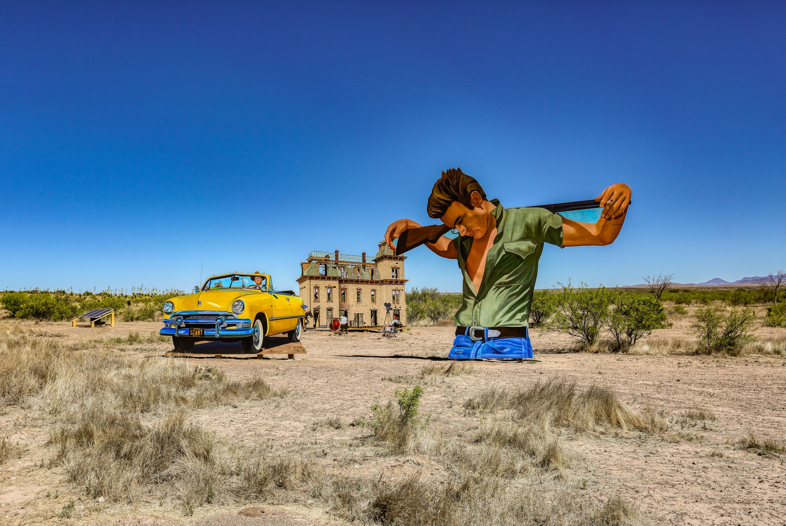 A giant art installation in Marfa, West Texas of a man with a green shirt and jeans holding a brown gun next to a yellow car and brown house. 