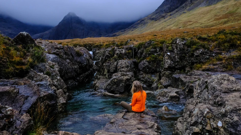 person wearing an orange sweatshirt sitting next to pool of water with mountains in the distance