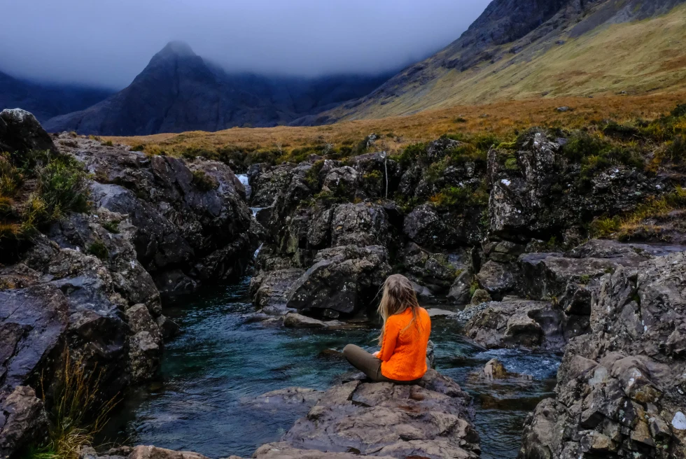 person wearing an orange sweatshirt sitting next to pool of water with mountains in the distance