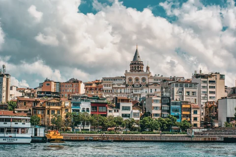City view of Istanbul across a river with white boats and blue red peach black and white colored buildings, green trees, and a white clouds with a blue sky