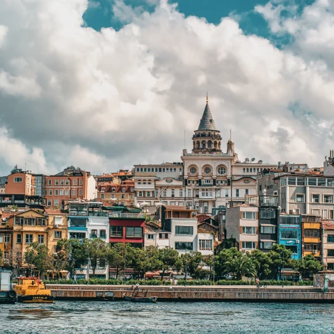 City view of Istanbul across a river with white boats and blue red peach black and white colored buildings, green trees, and a white clouds with a blue sky