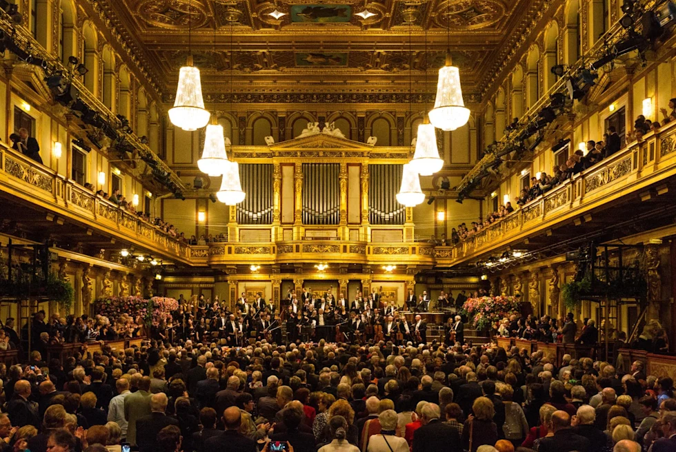 an ornate concert hall with gold details and large hanging light pictures at a sold out show with an orchestra