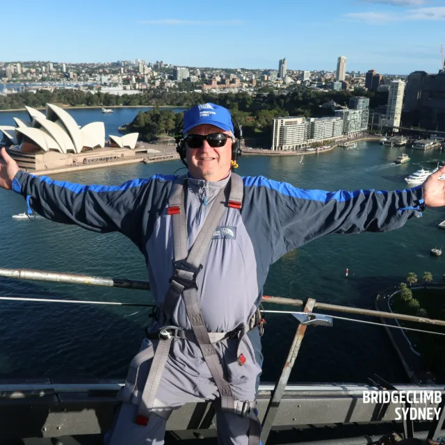 Picture of Ken at BridgeClimb Sydney wearing gear in front of a view of the Sydney harbor and opera house
