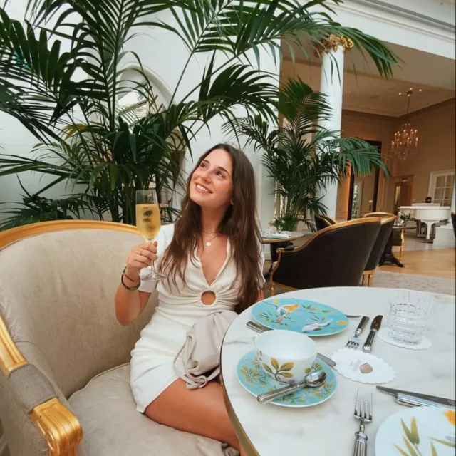 Travel advisor Sydney Alepa sits on a booth in a white dress and holds a glass of champagne