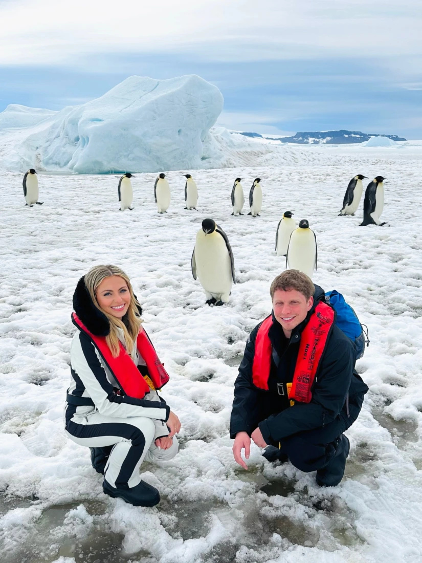Two people in the snow posing with a group of penguins