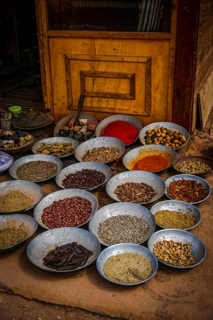 A spice market in Jordan with 20 silver bowls full of red, yellow, green and brown spices on a tan table with a brown wooden cabinet in the back.