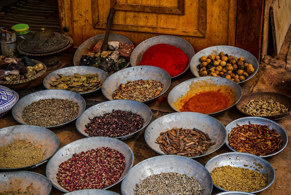 A spice market in Jordan with 20 silver bowls full of red, yellow, green and brown spices on a tan table with a brown wooden cabinet in the back.