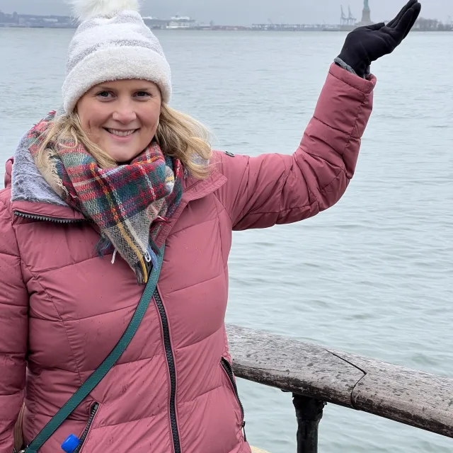 Travel advisor Lindsay Rice wears a pink coat, white winter hat and scarf and holds her hand up over a river
