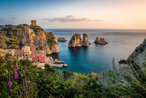 A spectacular view of a Sicilian coastal town, with natural cliffs bordering the sea, small houses and green grass, all under a soon-to-be setting sun.