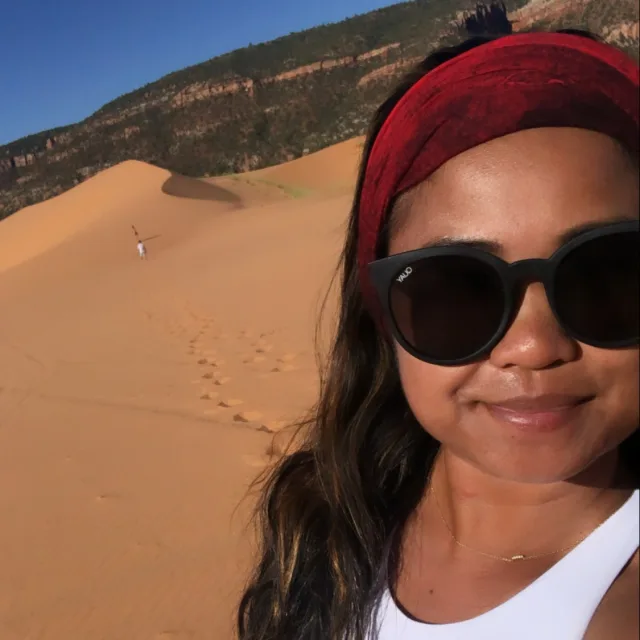 A selfie of Joanne in a white top and red headband wearing sunglasses and standing on a sand dune