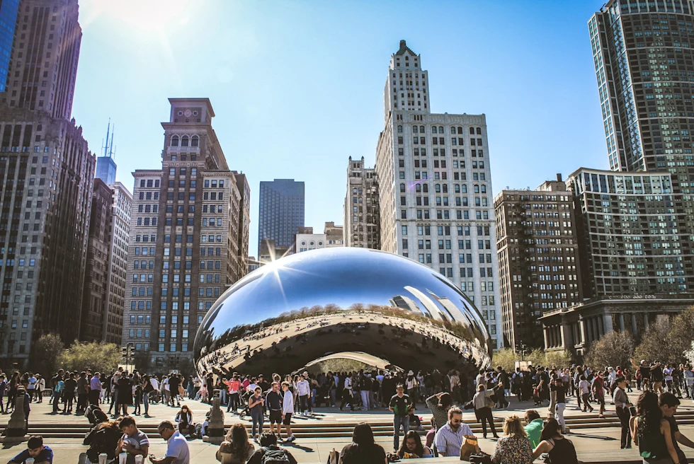 People walking around large silver structure with buildings in the background during daytime