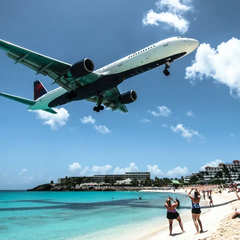 A white and blue passenger plane flying over a blue beach with white sand and tourists in St. Martin.  