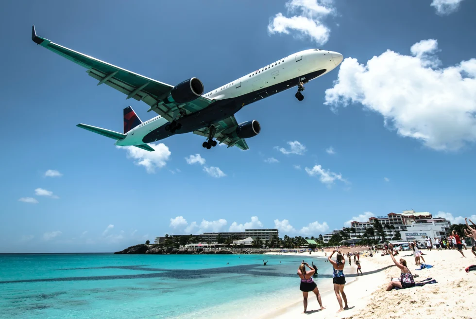 A white and blue passenger plane flying over a blue beach with white sand and tourists in St. Martin.  