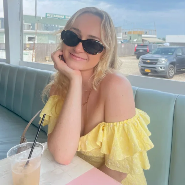 Emily Cameron in a yellow dress wearing black sunglasses and sitting on a sofa with a car in the background 