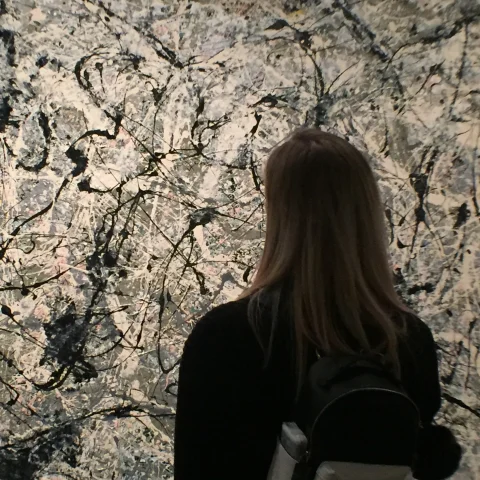 The back of a person with long brunette hair looking at a painting that has black, white, grey and cream splatters of paint all over it. 