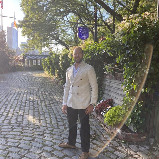 Fora travel agent Daniel Zarif wearing cream jacket and black pants standing next to green foliage on sunny day
