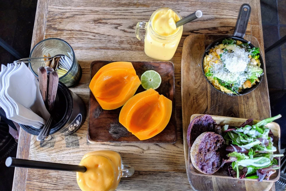 a fresh mango cut in half with half a lime, a yellow smoothie and side salads on a wood table