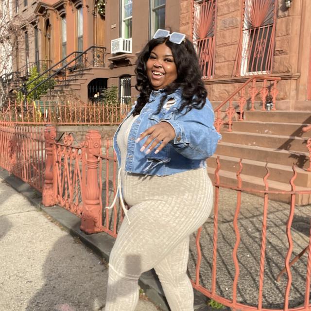 Woman wearing jean jacket and sunglasses poses in front of buildings.