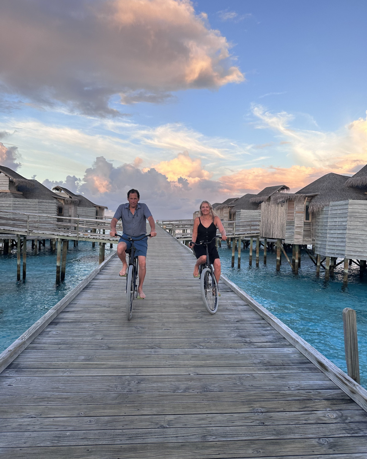 Two people cycling down a wooden bridge surrounded by beach bungalows and a beautiful cloudy sky. There is turquoise blue water on each side of the bridge too. 
