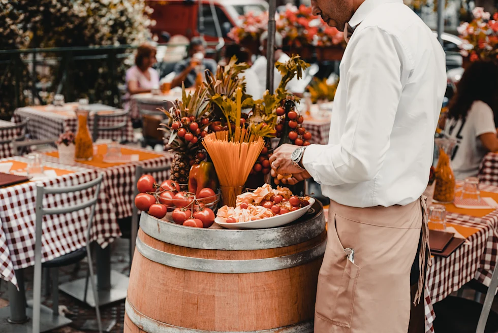 A host standing at a restaurant with a stand with pasta and vegetables, as well as tables that have checkered table clothes in Rome, Italy.