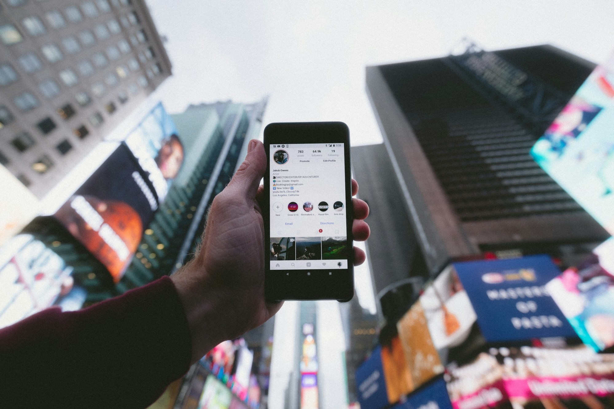 In Times Square, an unseen man holds his phone showing his Instagram profile before the skyline