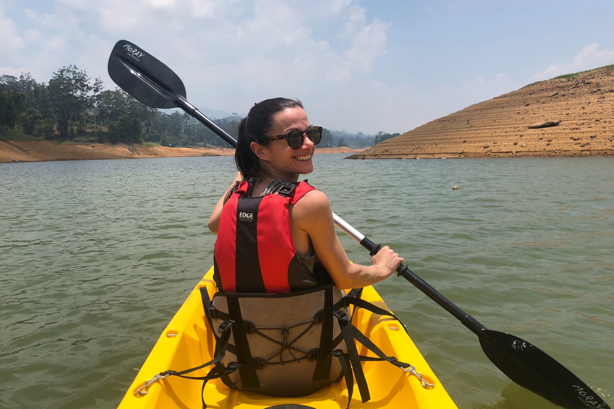 a woman wearing sunglasses in a yellow kayak and holding a black paddle looks back and smiles