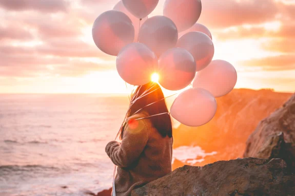Woman looking onto an ocean at sunset holding a handful of pink balloons.