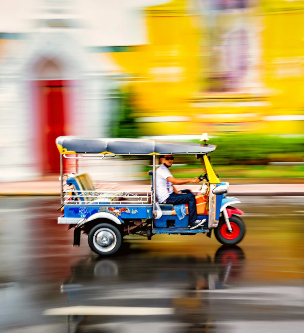 A man driving a tuk-tuk with an unfocused blurred colorful background of a street and yellow building