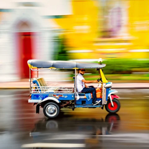 A man driving a tuk-tuk with an unfocused blurred colorful background of a street and yellow building