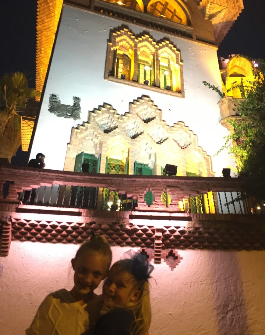 A view of two children smiling in front of a lit up building with intricate window frames, arches and white stone. 