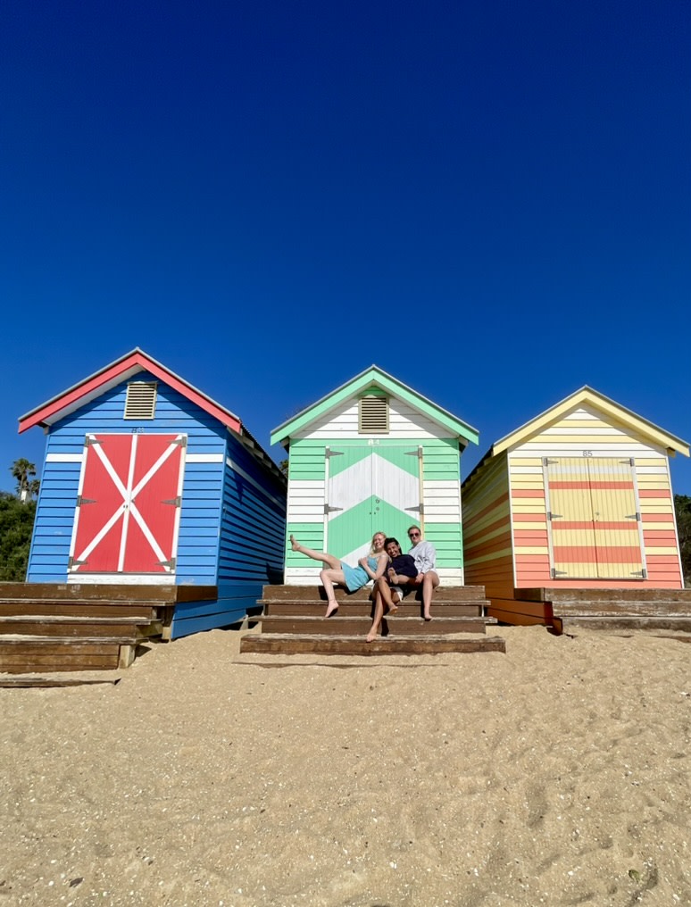 Small colorful beach houses on the beach with three people sitting on the front steps, against a bright blue sky
