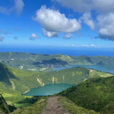 A dirt path leading to a view of a turquoise lake nestled into a green mountain range beneath the thick clouds and blue sky.  