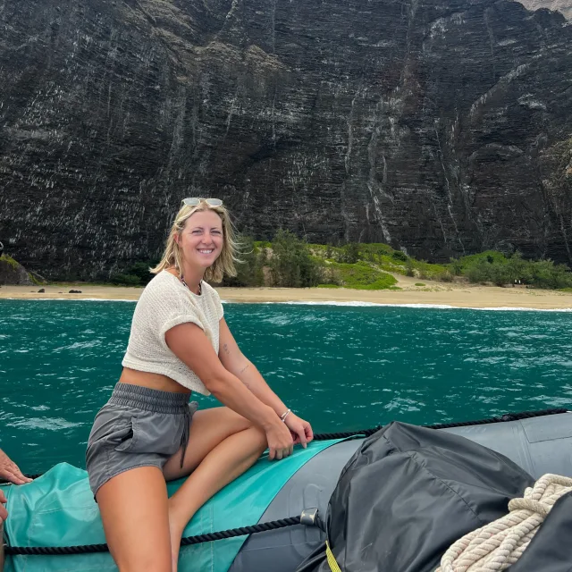 Samantha wearing a white crop top and athletic shorts on a boat with turquoise water and a rocky mountainside in the background