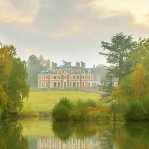 A view of Heckfield Place, a large red building with green roofs, in the distance surrounded by green grass, trees and a reflective body of water. 