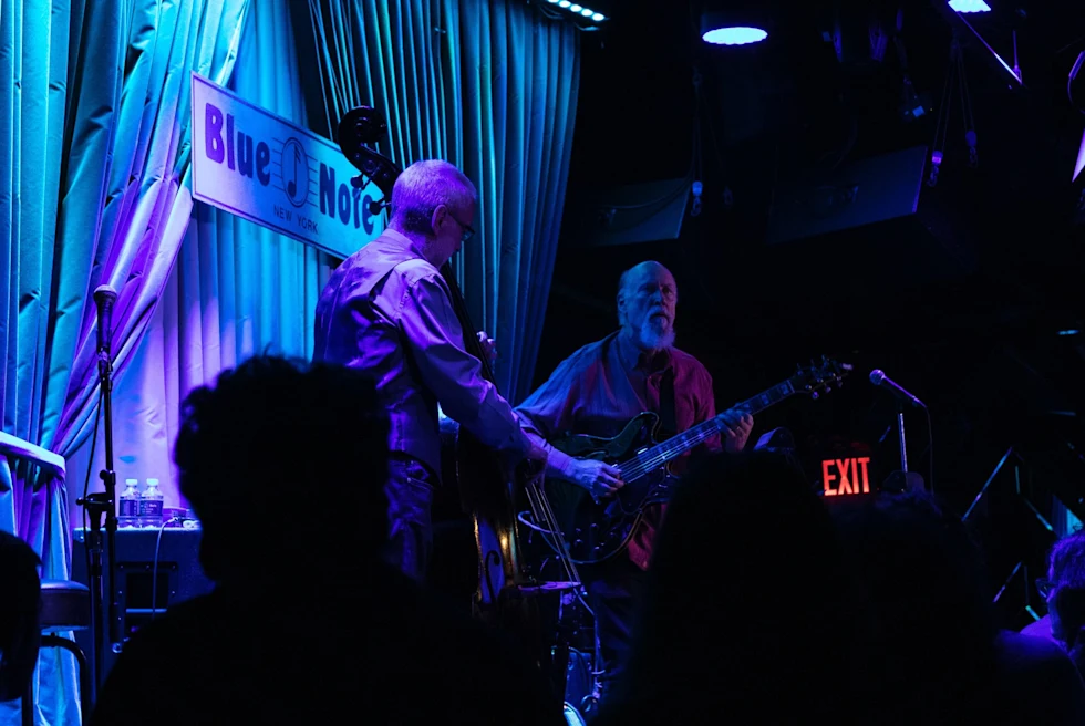 two men play instruments on stage in a dimly lit room with blue lighting