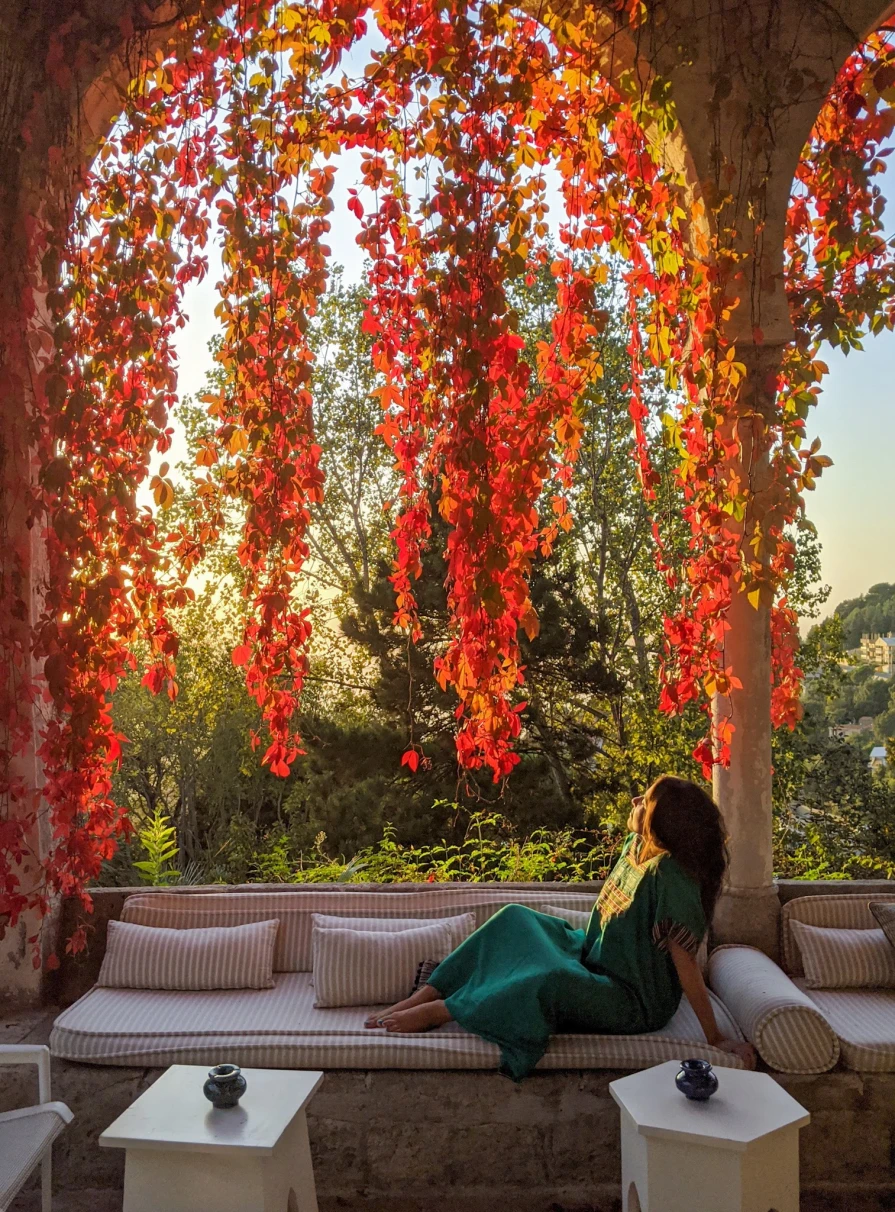 A girl in a dress sitting on an outdoor sofa framed by red leaves at sunset. 