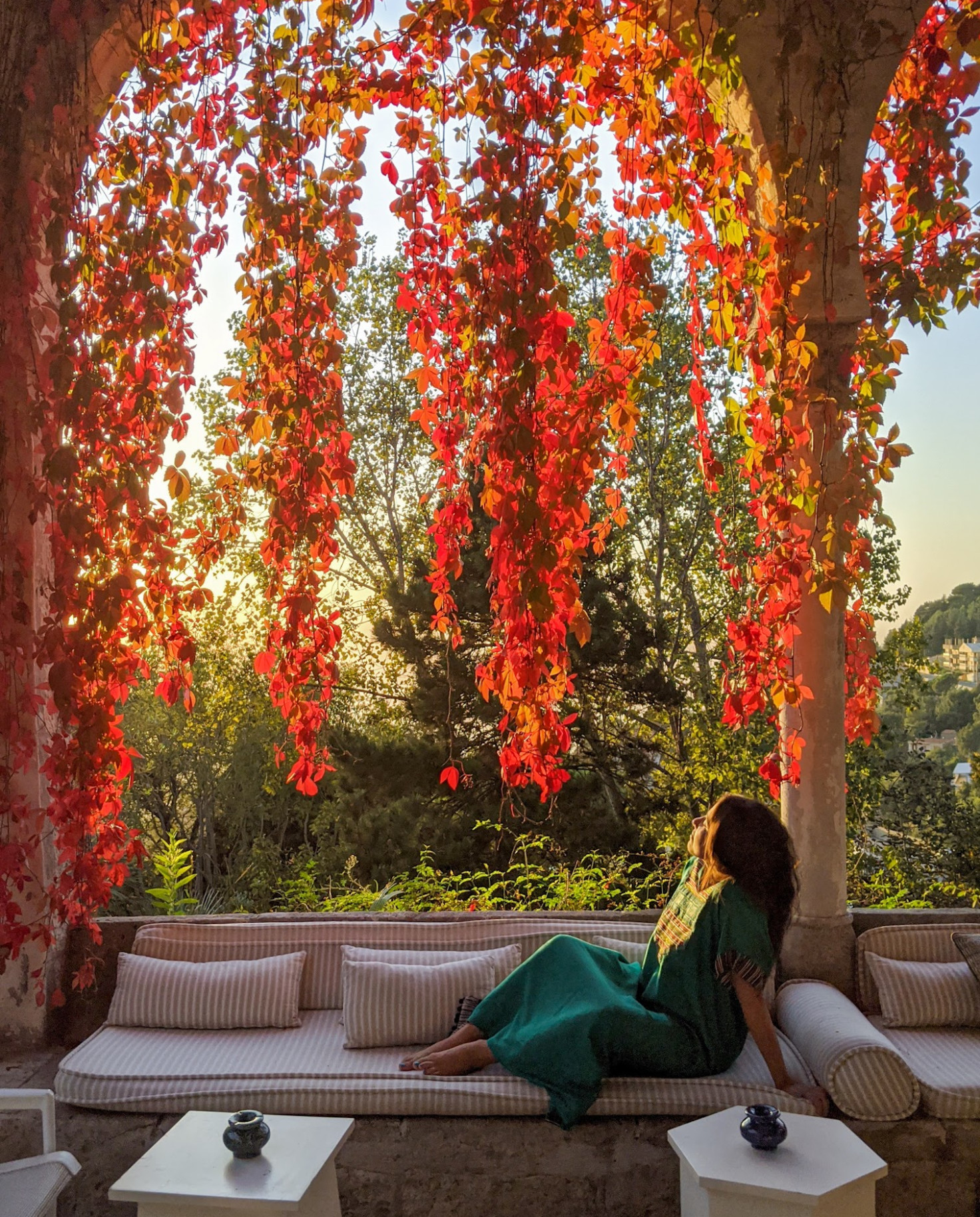 A girl in a dress sitting on an outdoor sofa framed by red leaves at sunset. 