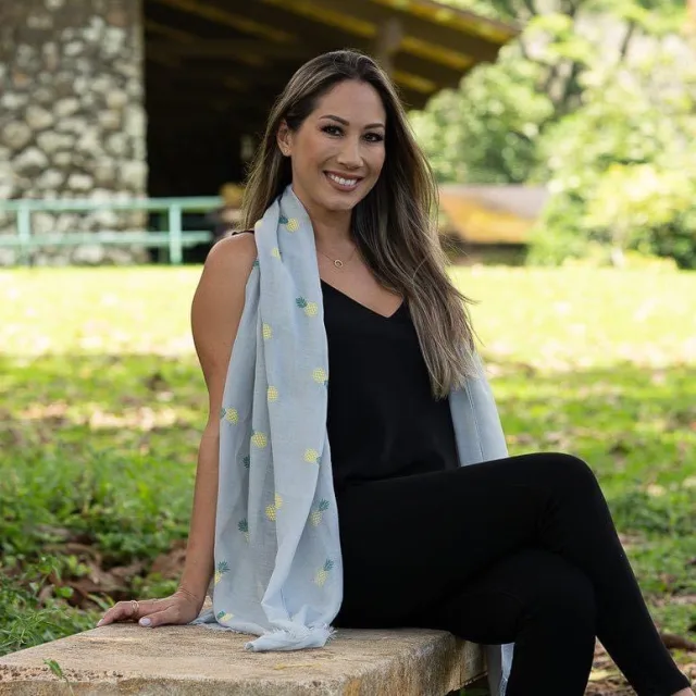 Elyssa wearing a black dress and blue scarf seated on a rock outside with green grass and trees in the background